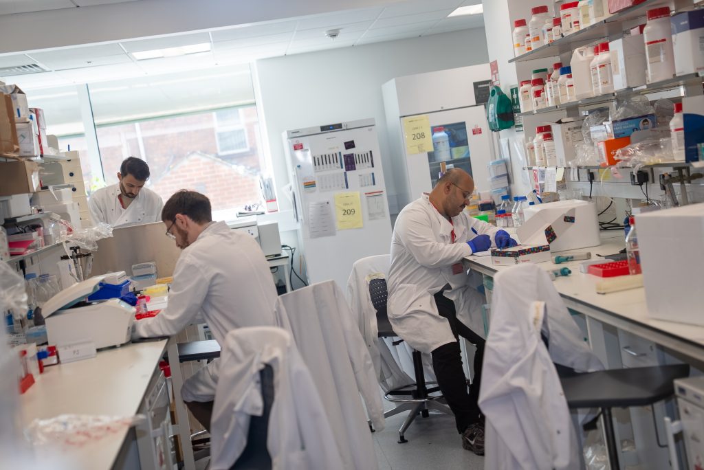 Photo of 3 colleagues working at benches in a lab environment. They are all wearing white coats and PPE gloves