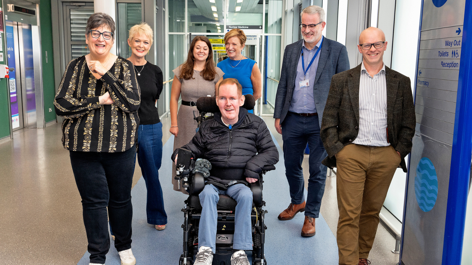Group of people walking together along a modern indoor corridor, including an individual using a powered wheelchair. The setting features glass walls, signage, and bright overhead lighting.