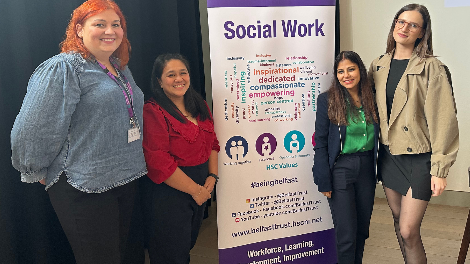 Four people standing beside a vertical banner that reads ‘Social Work’ with the Belfast Health and Social Care Trust logo at the top. The banner includes phrases such as ‘inspirational,’ ‘dedicated,’ ‘compassionate,’ and ‘empowering,’ along with HSC values icons and the hashtag #beingbelfast.